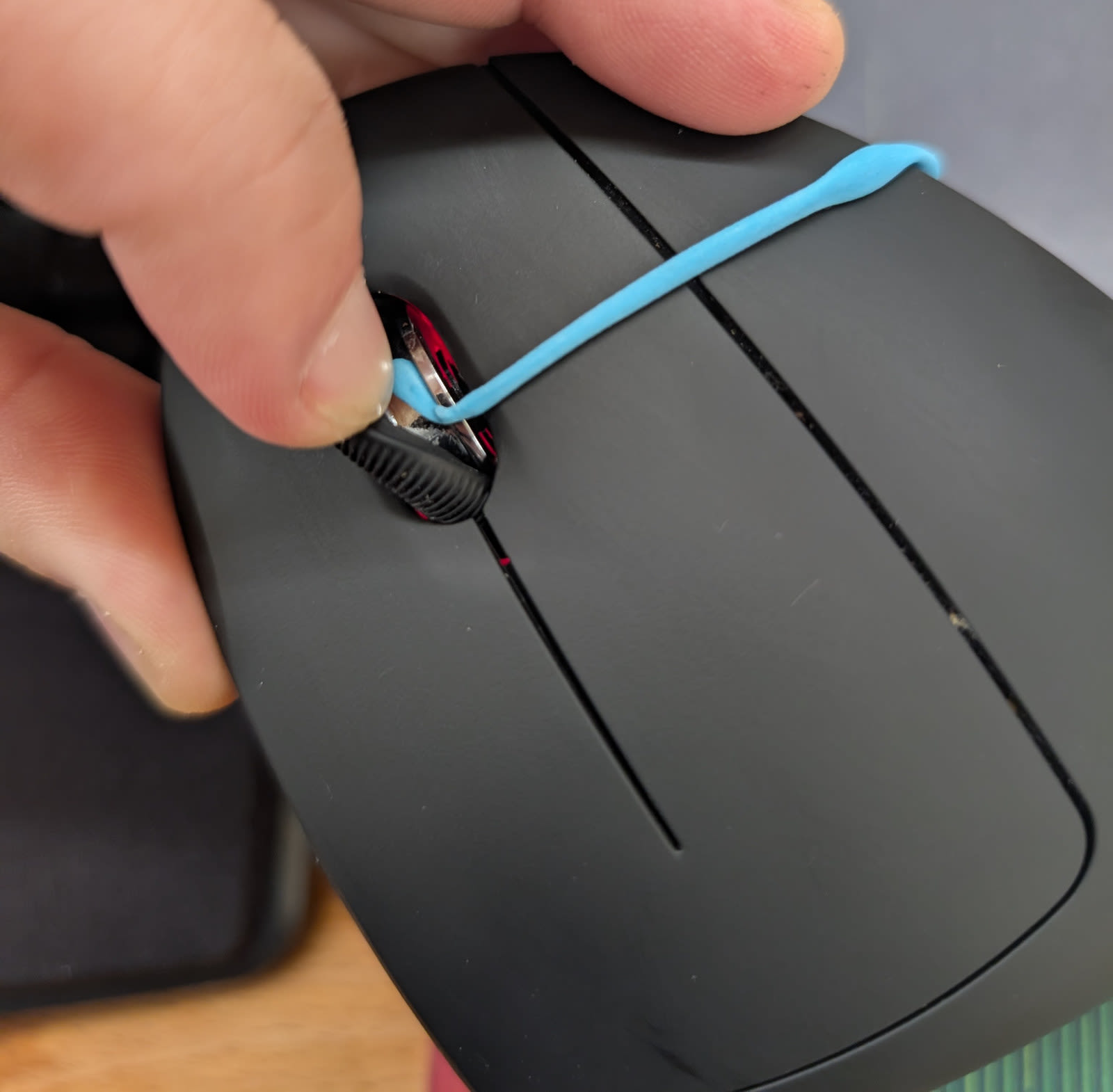 Close-up of two fingers holding the black vertical mouse. The thumb and index finger are lifting the black rubber wheel cover to reveal the silver ridged metal scroll wheel underneath. A thin string of bright blue adhesive putty is being inserted into the gap between the rubber cover and the metal wheel.