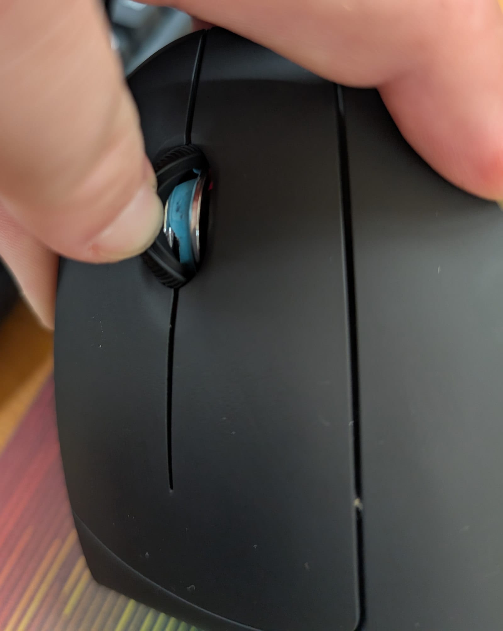 Close-up of the completed repair showing the black rubber scroll wheel with a hand touching it. Bright blue putty is visible in the channel between the black rubber wheel cover and the mouse body, filling the gap all the way around the wheel.