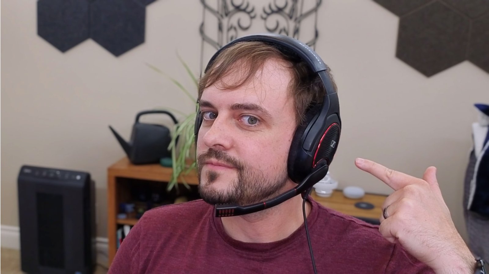 A well-lit portrait showing a person wearing a black Sennheiser gaming headset with red accent rings and an adjustable boom microphone, pointing at the headset with their finger. The image demonstrates clear, natural skin tones and good exposure.