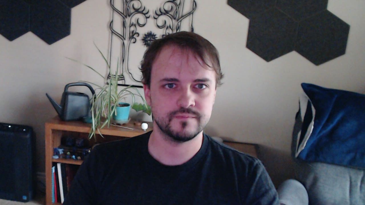 Logitech C920 webcam capture showing a person in very poor lighting conditions. The face is heavily shadowed and barely visible, the image is blurry with visible noise, skin tones appear unnaturally pink/purple, and background details are almost completely obscured in darkness.