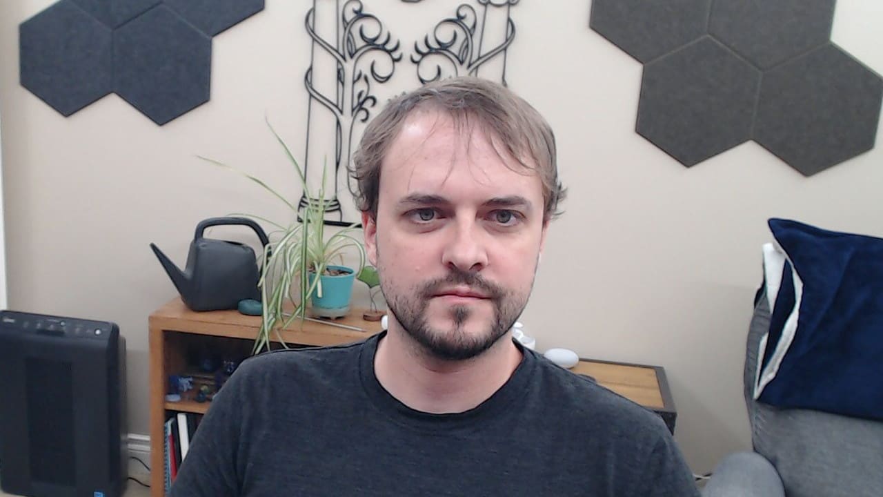 Logitech C920 webcam capture with bright overhead ceiling lights. The face is visible with more natural skin tones compared to the dark room shot, but pronounced shadows remain under the eyes, nose, and chin, creating an unflattering appearance.