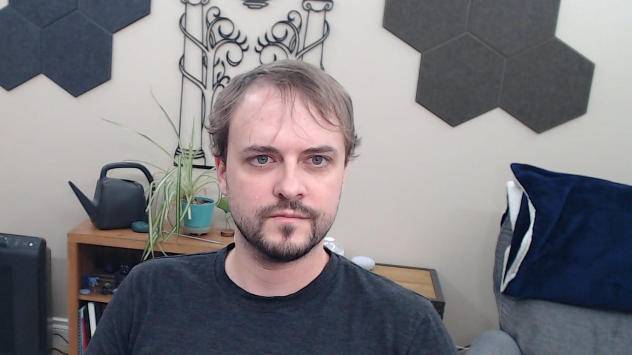 Logitech C920 webcam capture combining overhead ceiling lights with frontal lighting. The face is evenly illuminated with natural skin tones, minimal shadowing around the eyes and facial features, and significantly improved overall appearance compared to overhead-only lighting.