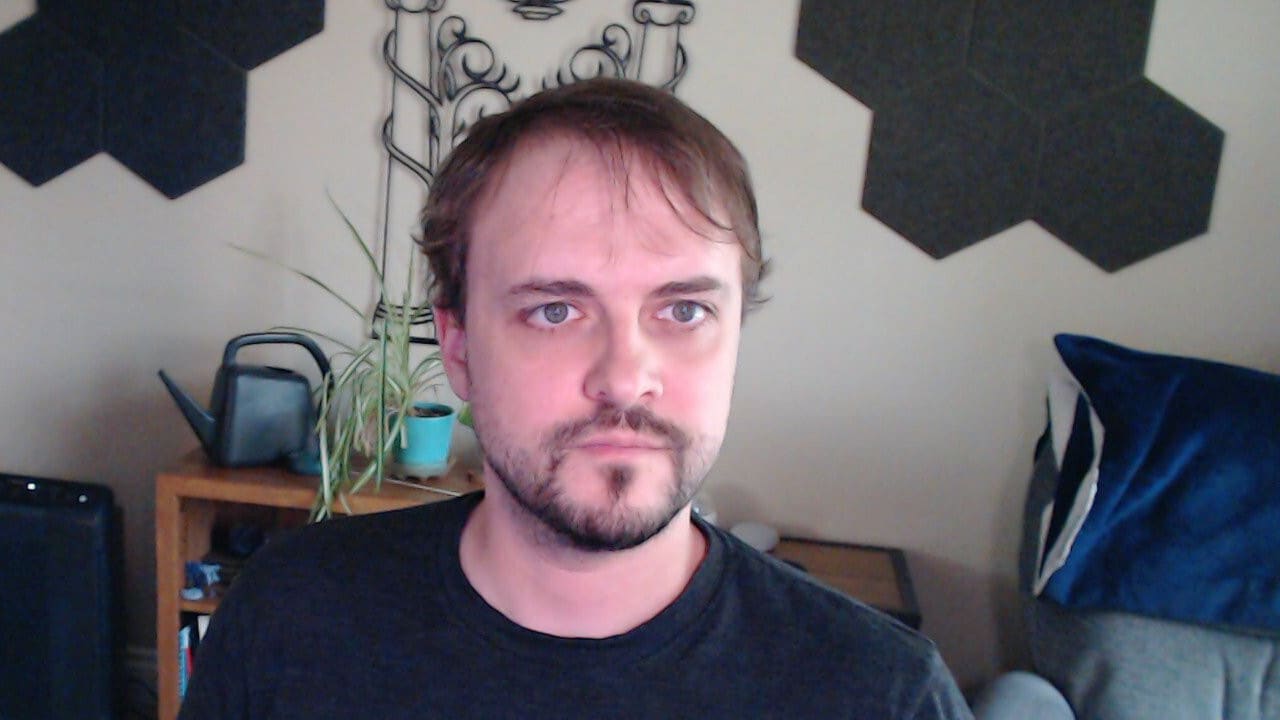 Logitech C920 webcam capture using only a monitor-mounted key light with minimal room lighting. The face is well-lit and evenly illuminated with natural skin tones and no harsh shadows, demonstrating that a single properly-positioned light can produce good results.